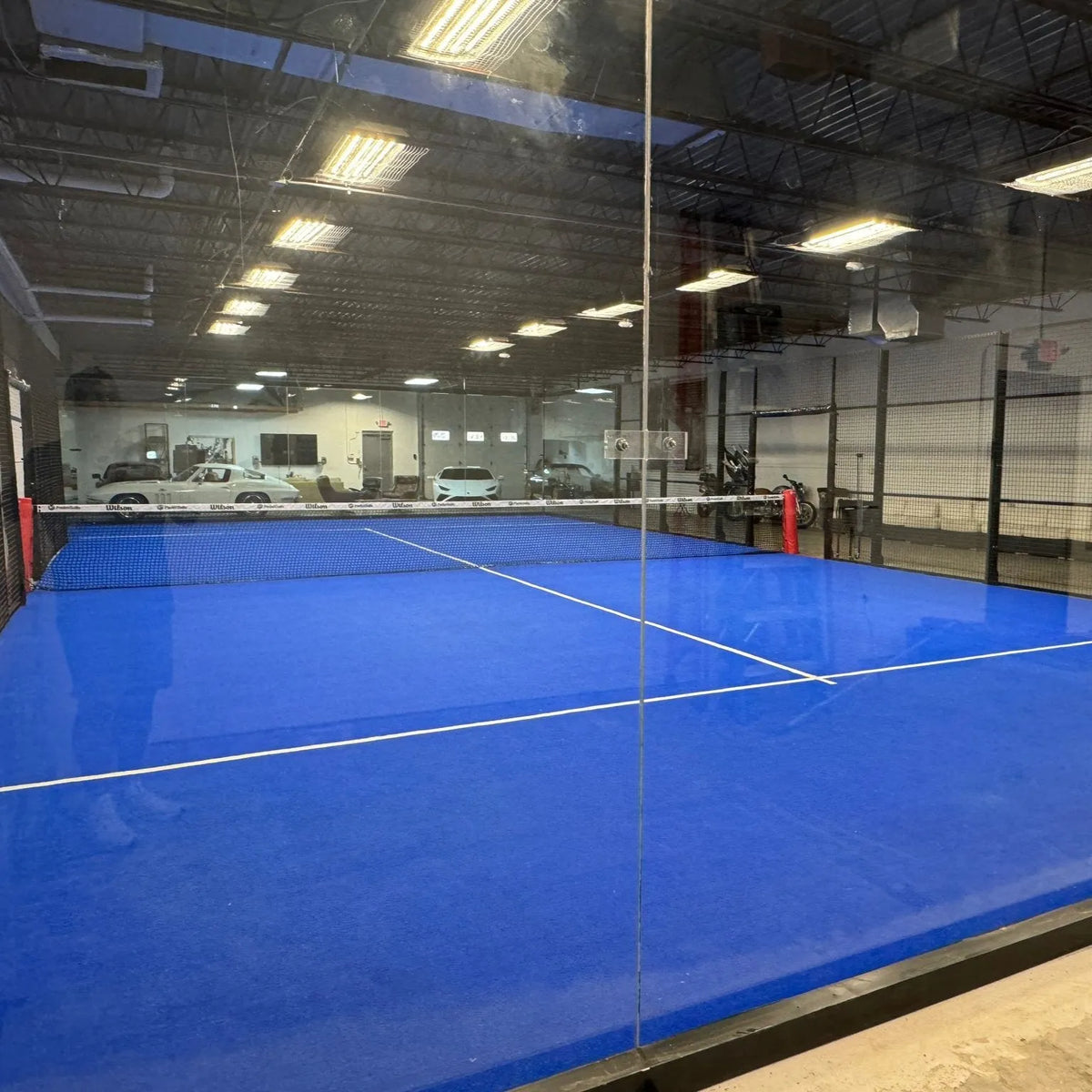 The Padel Smash indoor court rental offers bright blue flooring and white lines, enclosed by glass walls with a central net. Overhead, fluorescent lights brighten the scene surrounded by high black fences in an industrial setting, perfect for a 90-minute session per player.