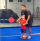 On a blue indoor sports court, a man assists a young boy holding a red paddle during a Padel Smash clinic. They focus on the swing with covered objects and other sports gear visible in the background.